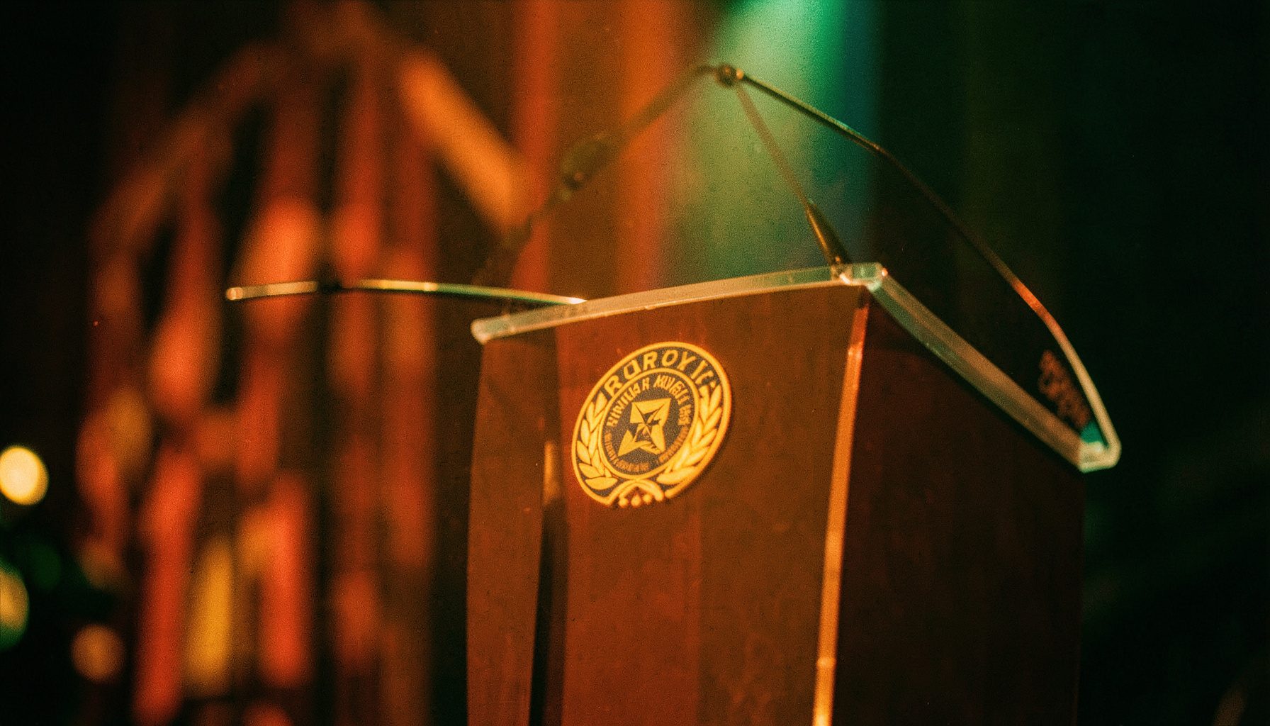 Warm cinematic view of a softly lit awards podium with a golden Rotary emblem, shallow depth of field, amber and green tones